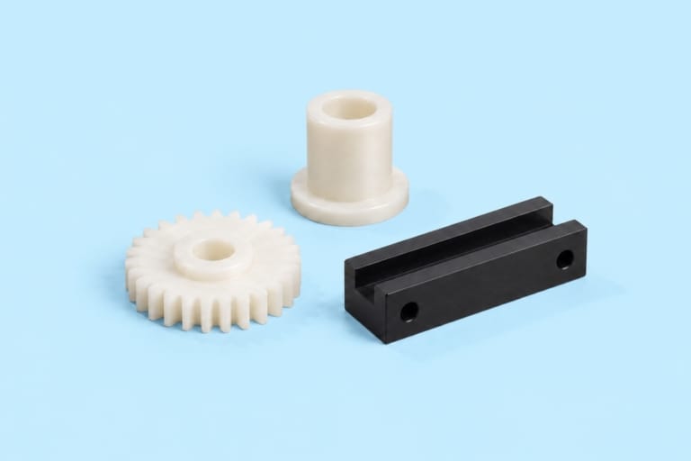 Vitt kugghjul, vit bussning och svart styrdetalj som illustrerar komponenter i POM-plast (acetal) mot ljusblå bakgrund.